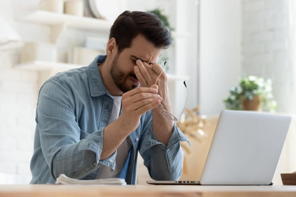 man wincing and rubbing eyes in front of laptop